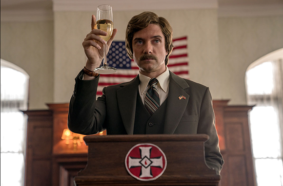 A man with a mustache in a suit raises a glass of champagne behind a wooden podium with a red cross emblem.