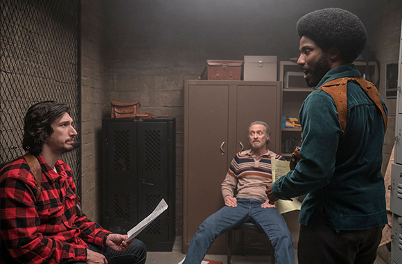 Three men in a dimly lit room; one sits in red flannel, one in a striped shirt, and one stands in a teal shirt.