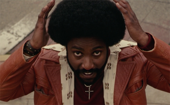 A man with a large afro and beard looks upward with his hands raised to his head.