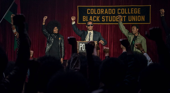 Three people stand on a stage with raised fists before a Colorado College Black Student Union banner.