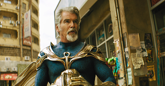 A man with grey hair and a beard wearing ornate gold and blue superhero armor in an urban setting.