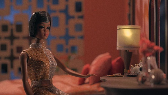 A fashion doll in a gold sequined dress sits in a dimly lit room next to a small lamp.