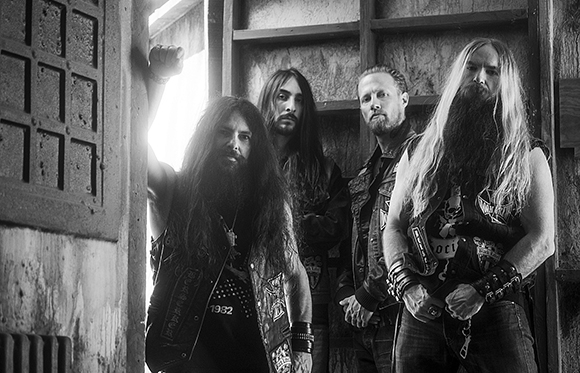 Four long-haired men in heavy metal attire posing together in a dark, rustic doorway.