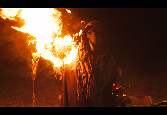A person wearing a dark, tattered hooded cloak breathes a large plume of bright orange fire.