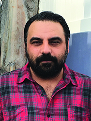 A man with dark hair and a thick beard wearing a pink and black plaid shirt.
