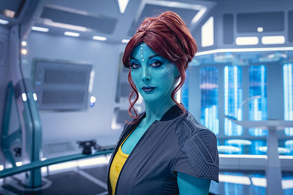 A blue-skinned woman with red hair and a gem on her forehead stands in a bright, futuristic sci-fi room.