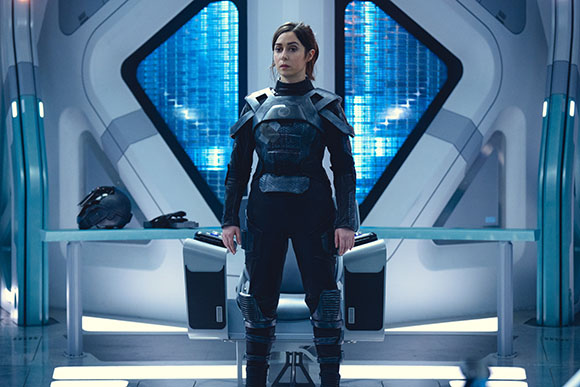 A woman in black tactical armor stands in a bright, futuristic white and blue corridor.