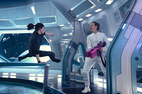 A woman in dark clothing lunges toward a man in white holding a purple device inside a spaceship.