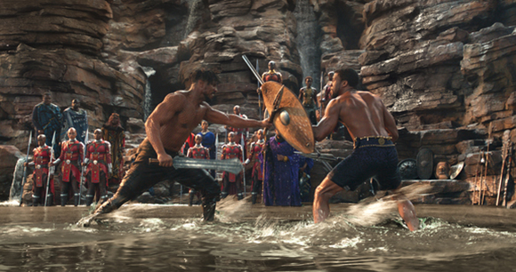 Two shirtless men duel with a sword and shield in shallow water against a rocky backdrop.