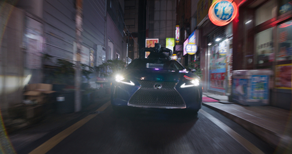 A dark car with bright headlights driving down a narrow, neon-lit urban alleyway at night.