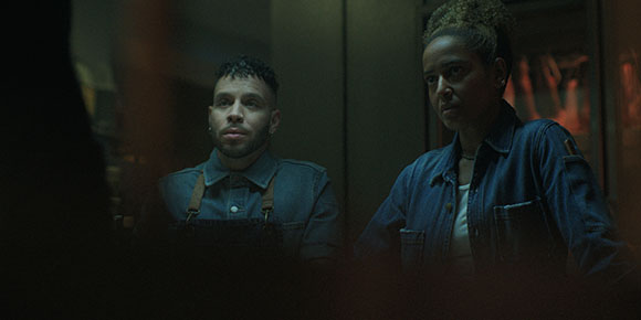 A man and a woman in denim workwear look intently toward something off-camera in a dark room.