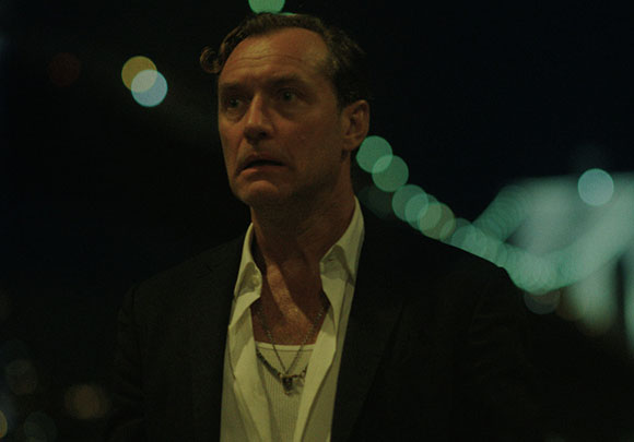 A man in a dark blazer and open white shirt looks startled against a dark background with bokeh lights.