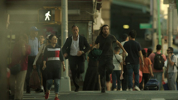 Two men run through a busy urban street filled with pedestrians during the day.