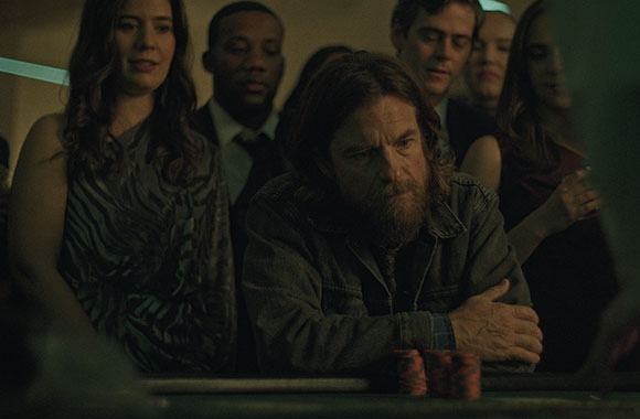 A man with a beard and long hair leans on a table while others watch him in a dark setting.