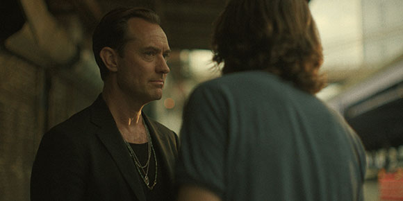 A man in a black suit and silver necklaces looks toward a person with long hair seen from behind.