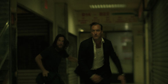 Two men running quickly through a dimly lit, narrow corridor.