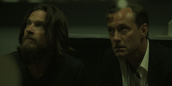 Two men with serious expressions look upward in a dark, moody environment.