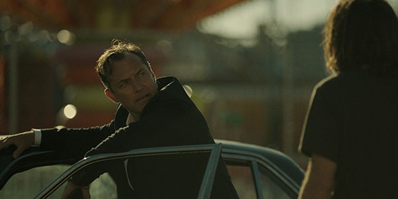 A man in a dark suit leans out of a car window, looking intensely at a person standing in the foreground.