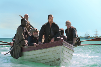 Five men in historical clothing row a small wooden boat across turquoise ocean water.