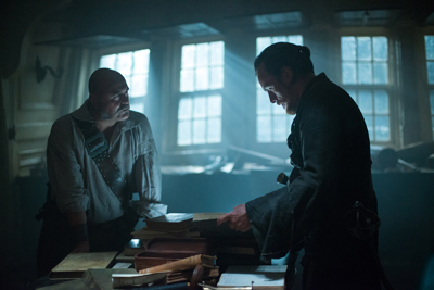 Two men in historical attire stand in a dark, sunlit cabin filled with books.