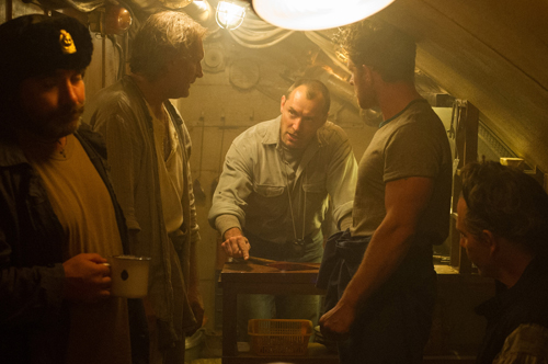 Five men stand in a dimly lit, rustic room, some looking down at a table.