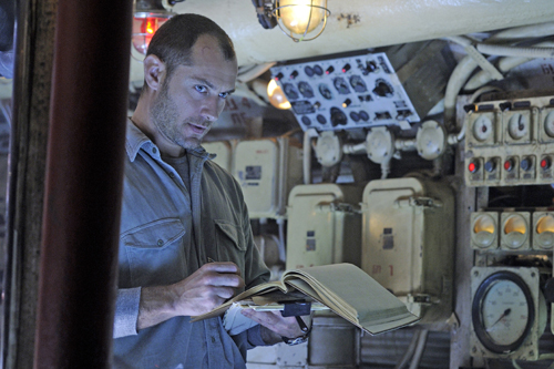 A man in a grey shirt holds an open book in a dimly lit, industrial room filled with machinery and wires.
