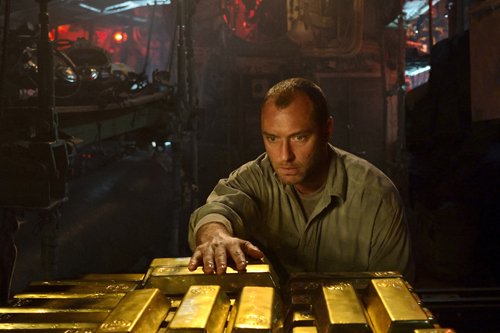 A man in a tan shirt leans over a collection of shiny gold bars in a dark, industrial room.