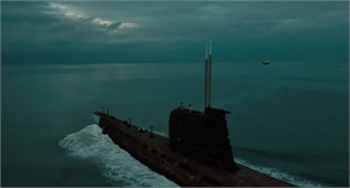 A dark submarine moves through choppy teal ocean water under a cloudy sky.
