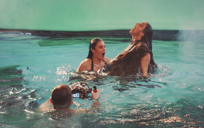 A camera operator films two women reacting dramatically in a pool against a green screen background.