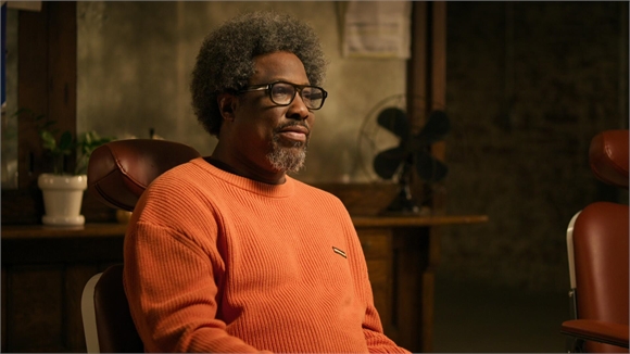 A man with graying afro hair and glasses sits in a chair wearing an orange sweater.