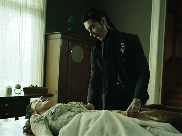 A man in a dark suit leans over a woman in a white lace dress lying on a table in a dimly lit room.