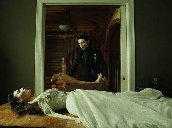 A man in a black suit stands behind a woman in a white gown lying on a wooden table.