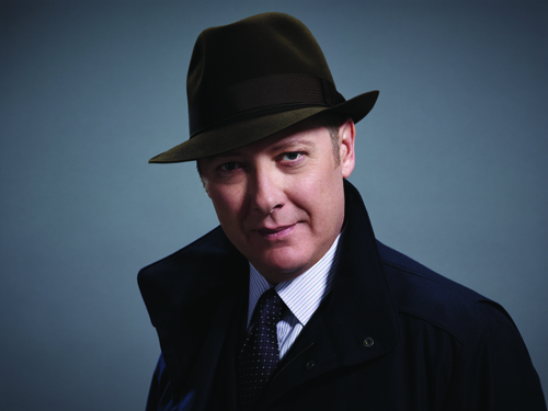 A man in a dark olive fedora and navy trench coat looking toward the camera.