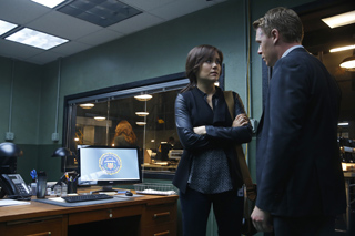 A woman with crossed arms looks at a man in a dark suit in an office setting.