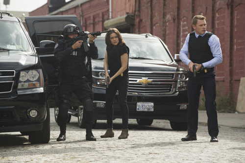 Three people in tactical gear stand near black SUVs on a paved street.