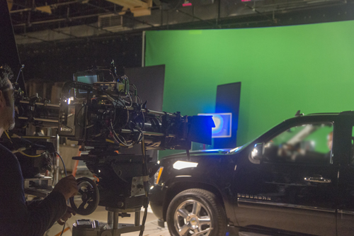 A professional film camera on a rig filming a black SUV in a studio with a large green screen background.
