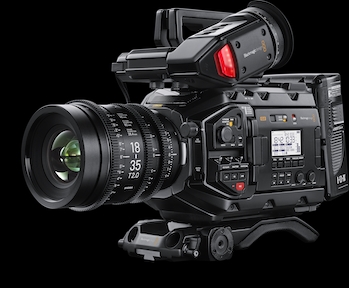A professional black video camera with a large lens and mounted viewfinder against a black background.