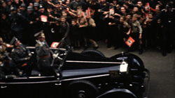 Military officers in a dark convertible drive past a large crowd of people waving flags.