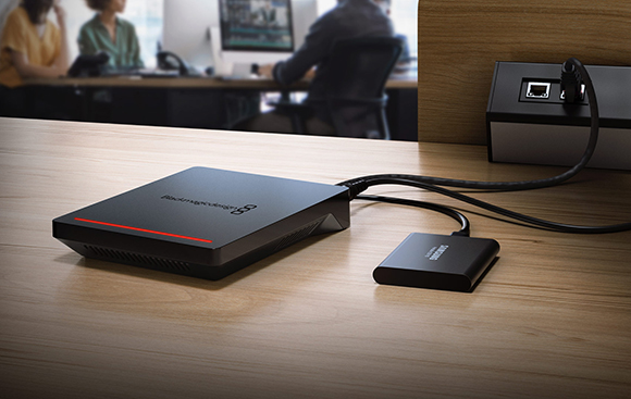 Black electronic devices and cables connected on a wooden desk in an office setting.