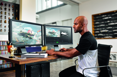 A man sits at a wooden desk using two large computer monitors displaying digital artwork.