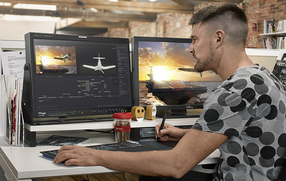 A man uses a stylus and tablet to edit digital artwork of an aircraft on dual computer monitors.