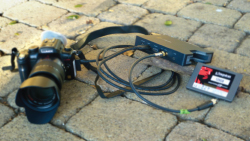 A black DSLR camera with a large lens connected by cables to a small black electronic device on cobblestones.
