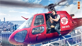 A cameraman sits in the open door of a red helicopter filming over a cityscape.