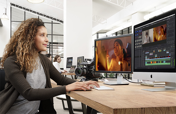 A woman with curly hair sits at a wooden desk using two large computer monitors to edit video footage.