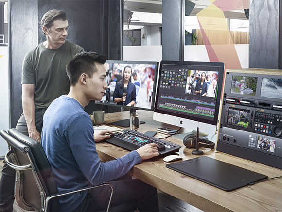 A man sits at a desk editing video on a computer while another man stands behind him watching.