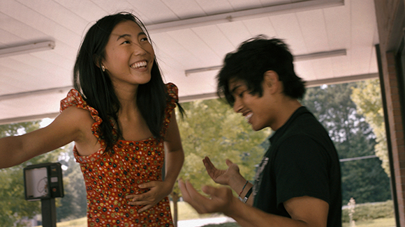 A smiling young woman in a red floral top and a young man in a black t-shirt laughing under a white awning.