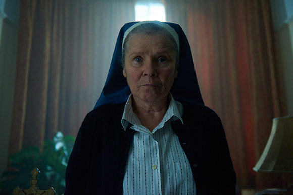 An older woman in a blue and white nun's habit looking forward with a serious expression.