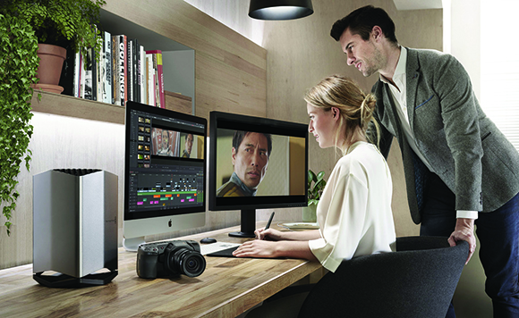 A man leans over a woman sitting at a desk with two computer monitors, a camera, and a silver computer tower.