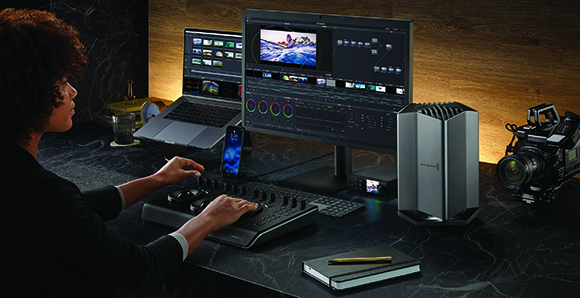 A person uses a control surface and large monitors to edit video in a dark studio.