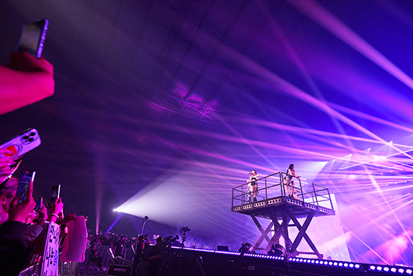 Two performers stand on a raised platform amidst bright purple and white stage lights at a concert.